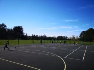A group of young pupils are pictured playing together inside a basketball court on a sunny day.