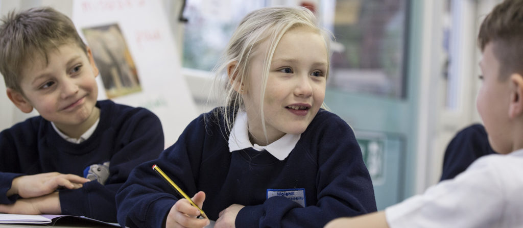 Three students seen sat next to each other at a table in a classroom, talking and smiling.