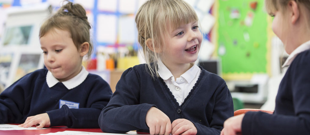 Three young female pupils are pictured sat together at a desk. Two are shown talking to each other, while the third is concentrating on her work.