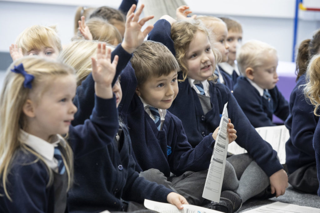 Students sitting on the floor with their legs crossed and hands up in the air to answer the teacher's question