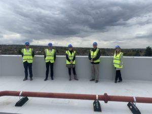 A group of five staff members are photographed standing on the rooftop of the Leigh Academy Rainham building, wearing hard hats and florescent coats.