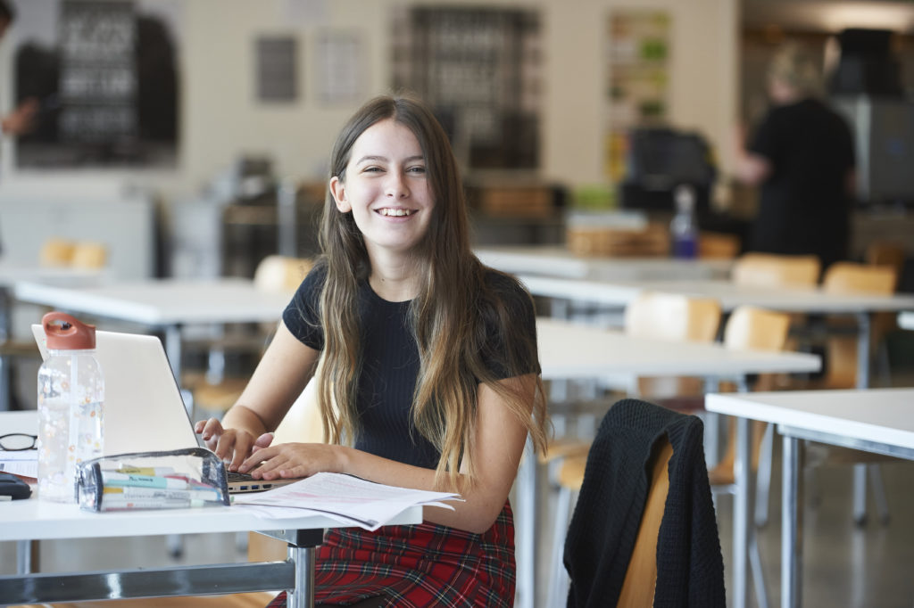 A sixth form student sitting on a table doing some artwork while smiling at the camera