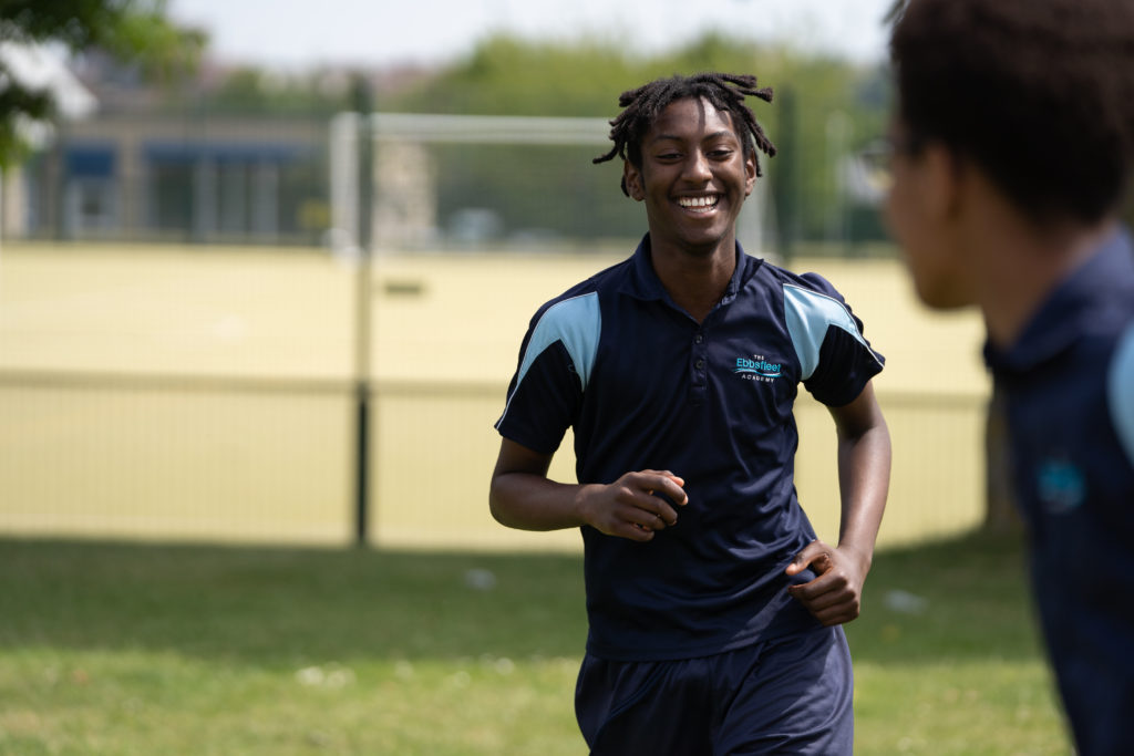 A male student seen dressed in an Ebbsfleet Academy PE Kit, is shown sprinting across a field and smiling, whilst playing a game of Football.