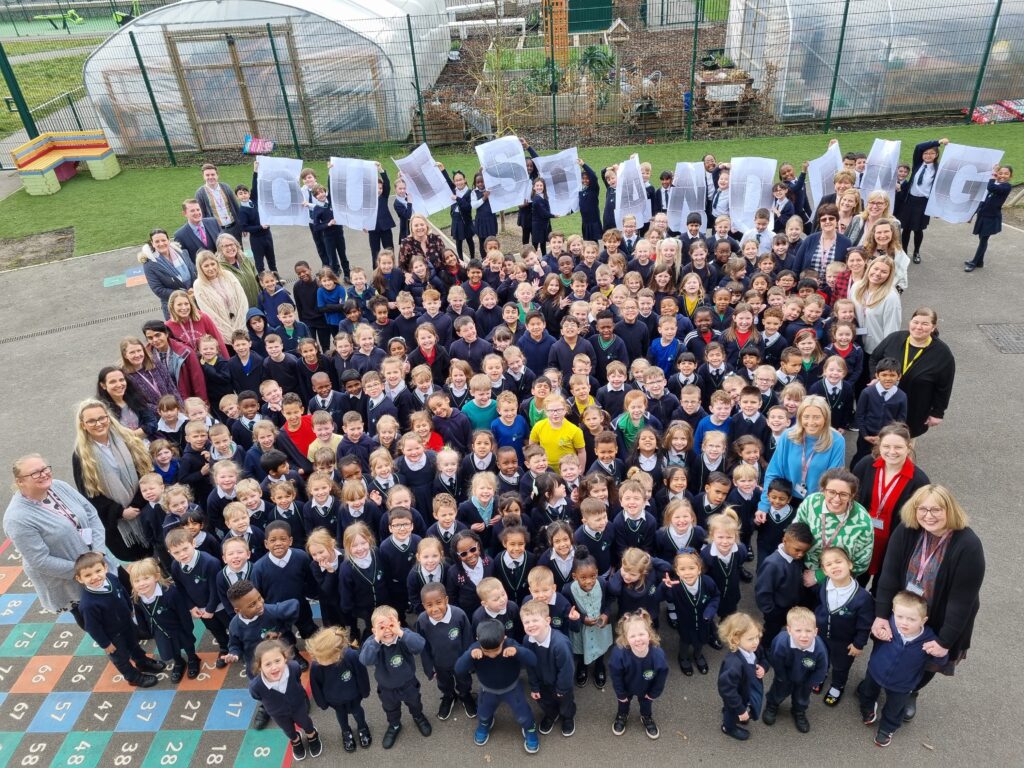 Many students in a group from Bearsted Primary Academy looking up at the camera smiling with staff and other students behind holding up paper that reads the word 'Outstanding'