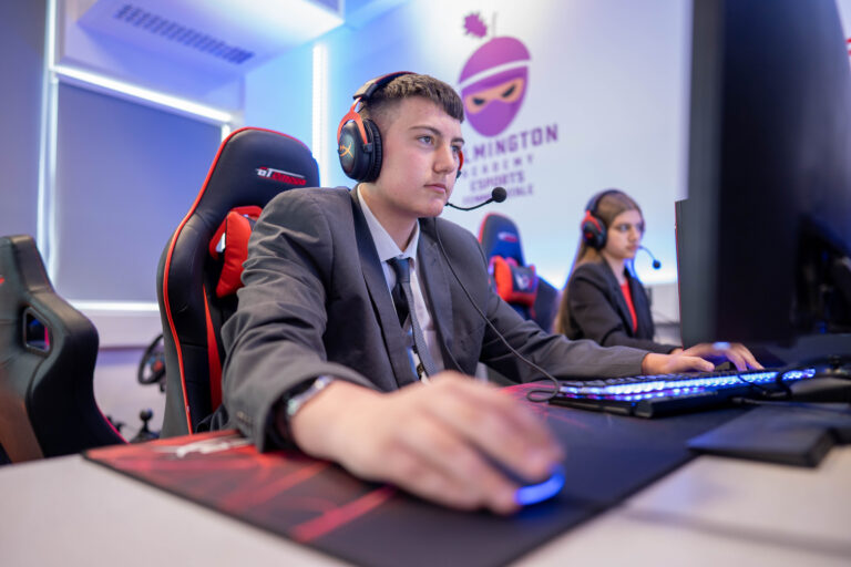 A post 16 student in a games classroom in front of a gaming pc with another student next to him.