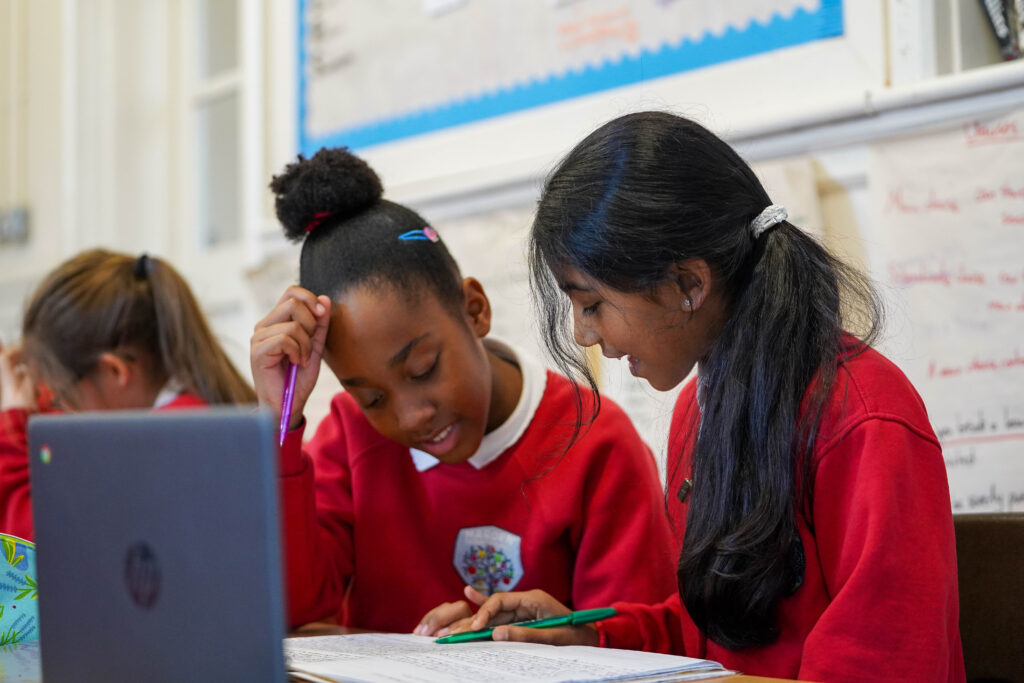Two girls in a class looking at a work book with a laptop in front of them.