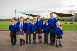 Students from various year groups are pictured huddled together for a group photo, outdoors on the academy grounds, smiling brightly.