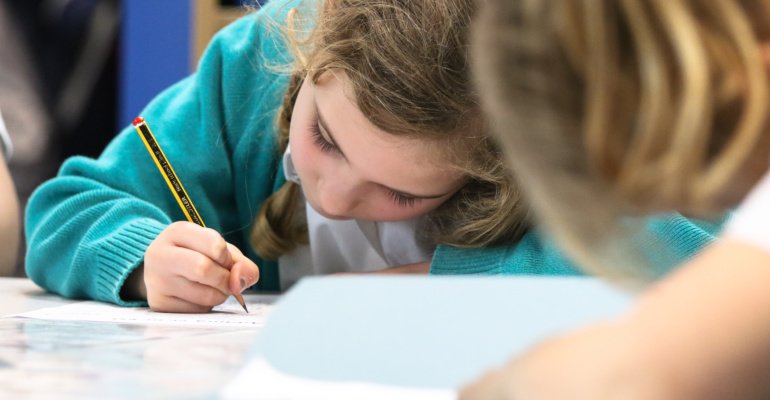 A young girl is seen sitting at her desk with her head down, focusing on a task, whilst writing with a pencil in her hand.