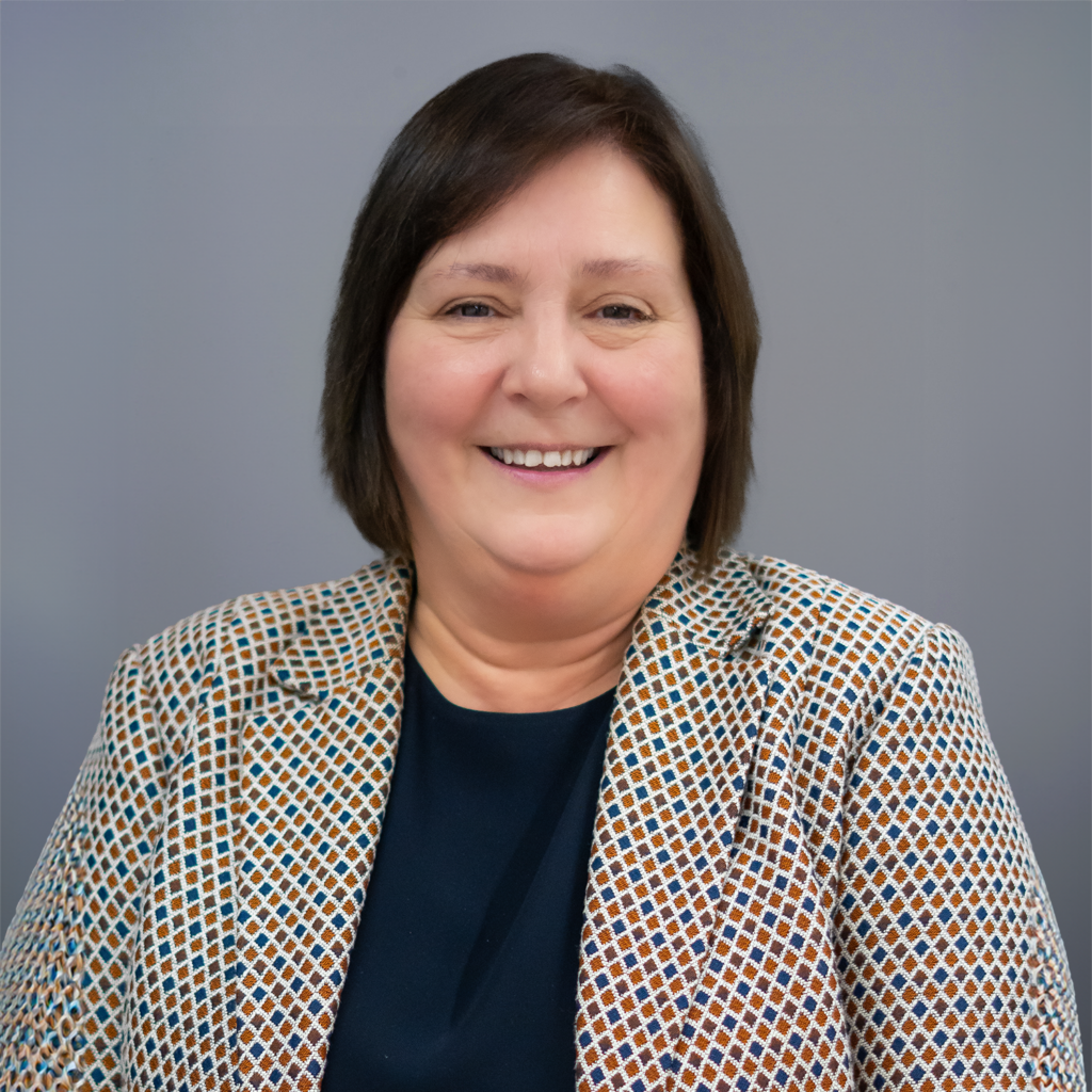 Head and shoulders portrait of a woman with short, dark brown hair smiling directly at the camera. She is wearing a blazer with a geometric pattern in brown, blue, and white over a dark top. The background is a solid gray.