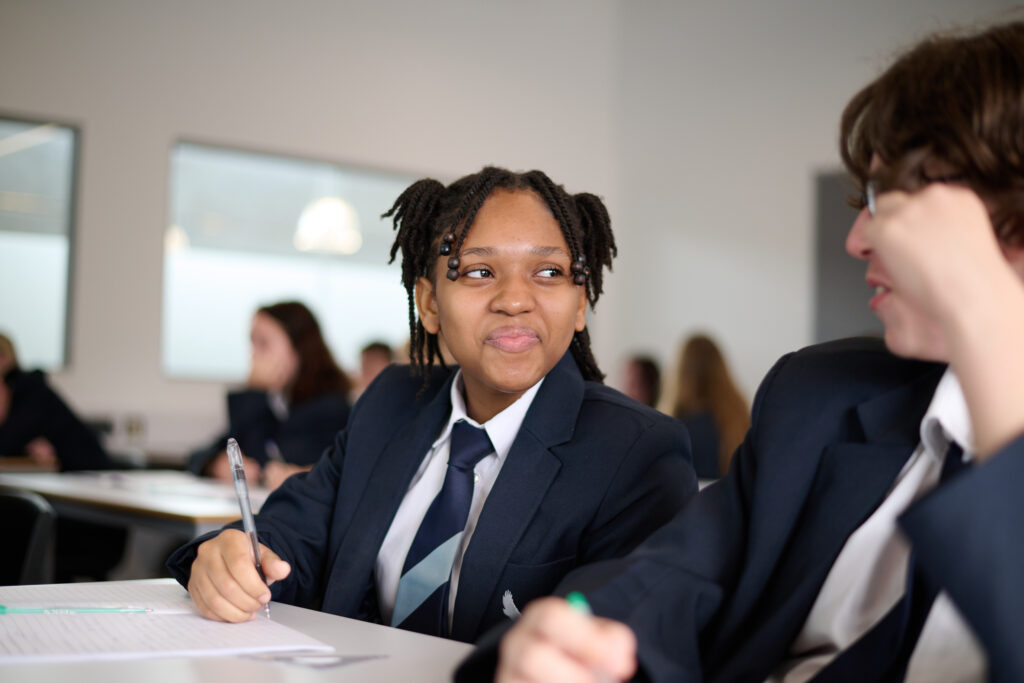 A medium shot shows two students in a classroom. On the left, a student with their hair in two braided ponytails, wearing a dark blue school blazer, white shirt, and striped tie, is holding a pen and looking to their right with a slight smile. The student on the right, partially visible with dark hair, also appears to be in a school uniform and is engaged in conversation. The classroom has light-colored walls and bright overhead lighting. Other students are blurred in the background.