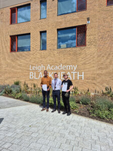 This photo shows students with an adult male (likely a teacher or staff member) in front of a building with "Leigh Academy BLACKHEATH" visible on its facade. The students are holding papers, suggesting they are receiving their results or official documents. The bright outdoor setting and the papers held by the students indicate an important day, often associated with exam results.