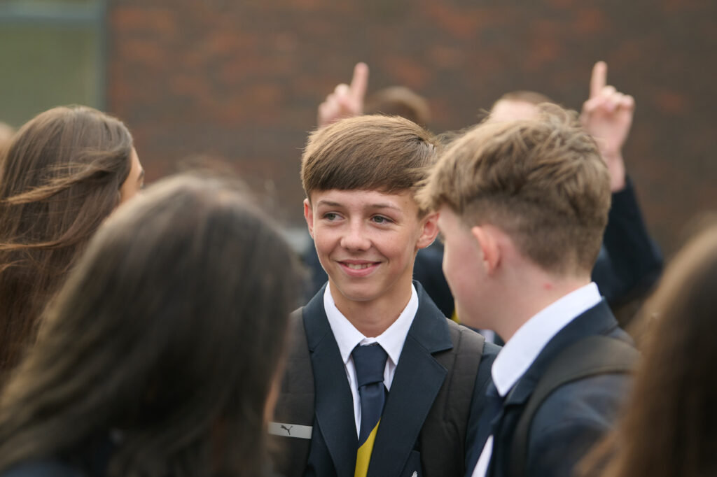 A close-up shot captures a young person with short, light brown hair, wearing a dark school blazer, white shirt, and dark tie, smiling and looking to their right. Another person's shoulder and the back of their head are visible in the foreground on the right. In the background, other blurred figures are present, with one person's hand raised, pointing upwards. The setting appears to be outdoors.
