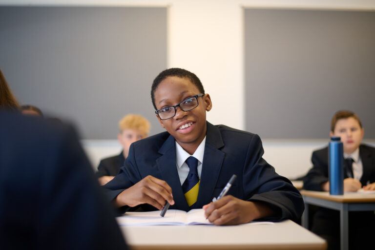 A student wearing glasses, a dark school blazer, white shirt, and dark tie with yellow accents, is seated at a desk in a classroom. The student is holding a pen over an open notebook and looking forward with a slight smile. The background is blurred, showing other students at their desks and a grey wall with a whiteboard.