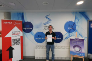 A young man is standing alone, holding white papers, and smiling. He is also in the same Strood Academy location.
