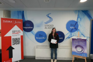 A young woman is standing alone, holding white papers, and looking towards the camera. The setting is the same Strood Academy environment.