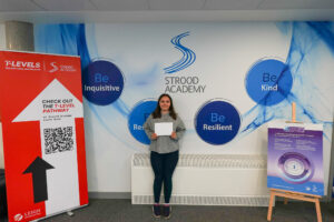 A young woman is standing alone, holding white papers, and smiling. The background is identical to the other photos, featuring the Strood Academy branding and the educational pathway information.