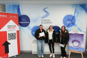 Three young women are standing side-by-side, each holding white papers, likely their results. They are in front of a wall with "STROOD ACADEMY" prominently displayed, along with positive attributes like "Be Inquisitive," "Be Kind," "Be Respectful," and "Be Resilient." There is a poster about "The IB Career-related Programme (CP)."