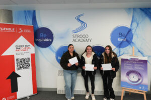 Three young women are standing side-by-side, each holding white papers, likely their results. They are in front of a wall with "STROOD ACADEMY" prominently displayed, along with positive attributes like "Be Inquisitive," "Be Kind," "Be Respectful," and "Be Resilient." There is a poster about "The IB Career-related Programme (CP)."