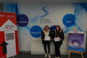 Two young women are holding up documents (likely results) in front of them. They are standing in the Strood Academy with the "Be Inquisitive" and "Be Respectful" circles visible behind them.