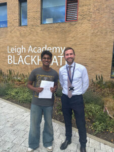 This photo shows a student with an adult male (likely a teacher or staff member) in front of a building with "Leigh Academy BLACKHEATH" visible on its facade. The students are holding papers, suggesting they are receiving their results or official documents. The bright outdoor setting and the papers held by the students indicate an important day, often associated with exam results.