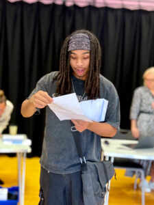 A young man with dreadlocks and a bandana is holding papers (likely results) and a white envelope. he's looking down at the papers in his hands with a surprised or focused expression.