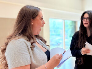 Two young women are interacting. the woman on the left has a surprised, open-mouthed expression while looking at her papers, and the woman on the right is looking at her.