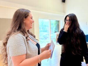 Two young women are interacting. the woman on the left is smiling and looking at the woman on the right, who is also smiling, suggesting a positive exchange about their results.