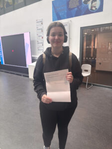 A young woman wearing headphones and carrying a backpack is holding a document that appears to be an "International Baccalaureate Organization" results sheet. She is smiling, likely pleased with her results.