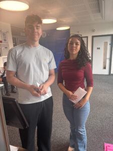 A young man and a young woman are standing together, both holding white papers or envelopes. They are smiling