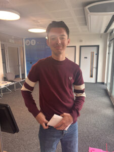 A young man is smiling at the camera while holding a white envelope tucked into his jeans. He appears happy and relaxed.