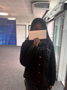 A young woman is holding a white envelope in front of her face, partially obscuring it, and smiling or reacting to what's on the envelope. The background has a blue wall with words like "thinkers," "Principled," and "Risk-takers," suggesting a school with a focus on character development.
