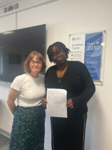 This photo shows a woman and a student, both smiling, holding a document inside a building. Behind them, a display titled "LEIGH Academy Excellence Charter Vision 2030" is visible, further indicating a school or academy setting. The student is holding papers, likely their exam results or another form of academic achievement.