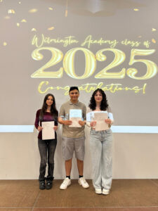Three students standing on the stage in front of the same "Wilmington Academy class of 2025 Congratulations!" display, proudly holding their certificates. They are likely posing for individual or small group portraits during the ceremony.