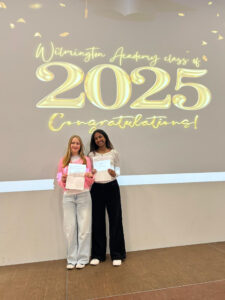 Two students standing on the stage in front of the same "Wilmington Academy class of 2025 Congratulations!" display, proudly holding their certificates. They are likely posing for individual or small group portraits during the ceremony.