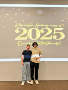 Two students standing on the stage in front of the same "Wilmington Academy class of 2025 Congratulations!" display, proudly holding their certificates. They are likely posing for individual or small group portraits during the ceremony.