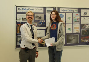 A man with glasses and a tie is shaking hands with a young woman. Both are smiling and the woman is holding some papers. They are standing in front of a display board with various posters and images