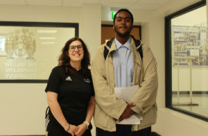 A woman with glasses and a black Adidas shirt is standing next to a young man. Both are smiling. The man is holding some papers. Behind them, there's a window with some text that appears to be reversed and a crest.