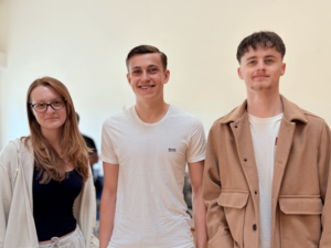 Three young adults are standing side-by-side, looking at the camera. The person on the left is a female with long, reddish-brown hair and glasses, wearing a light-colored cardigan over a dark top. The person in the middle is a male with medium-length brown hair and a beard, wearing a brown t-shirt with "WEST POINT" written on it. The person on the right is a male with short, dark hair, wearing a light-colored t-shirt under a light brown jacket. They appear to be indoors against a light-colored wall.