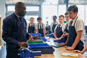 A teacher with a chisel in his hands is talking to six pupils, wearing blue aprons, over their school uniform during a class. There is a wooden bench and various other tools on show.
