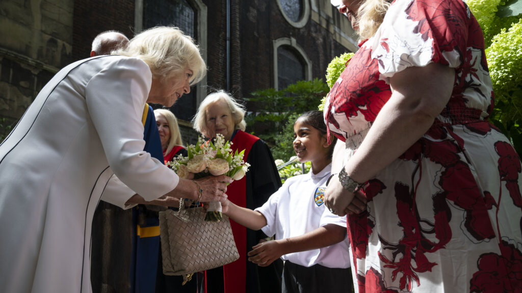 Queen Camilla, wearing a white jacket, is receiving a bouquet of flowers from a young girl. Another woman in a red and white patterned dress and a woman in academic regalia are also present. The scene appears to be outdoors, with a building and greenery in the background.