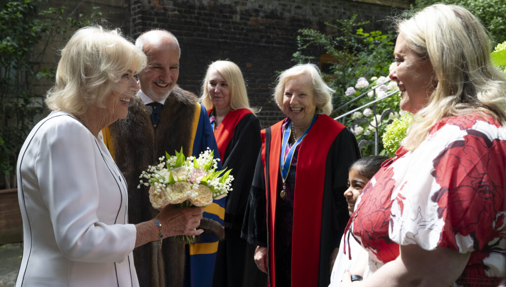 Queen Camilla, on the left, is looking towards and possibly speaking with the woman in the red and white floral dress, who is smiling. Queen Camilla is still holding the bouquet of flowers. Between Queen Camilla and the woman in the floral dress, a man in a fur-trimmed robe and a woman in red academic-style robes are visible, both smiling and looking towards the interaction. The young person in the school uniform is also visible on the far right, smiling and looking in the same direction. The background is similar to the first image, with a brick building and green foliage, suggesting it was taken in the same outdoor location.