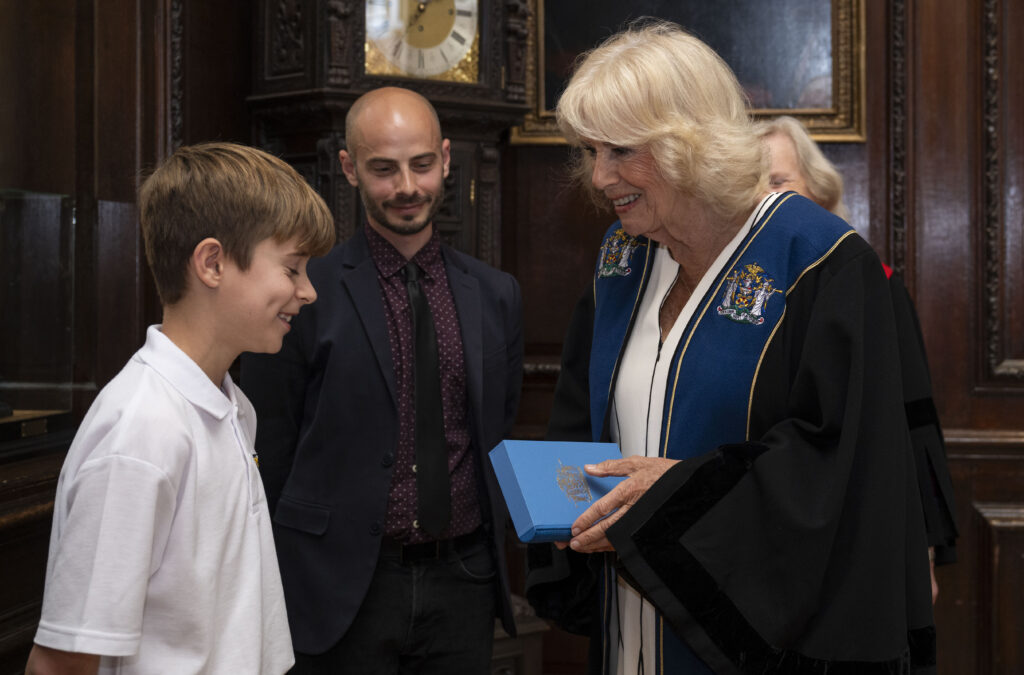 Queen Camilla, wearing academic regalia, is presented with a blue box from a young boy. Both are smiling. A man is standing behind the boy, also observing. The background features dark wood paneling, a grandfather clock, and framed portraits, suggesting an indoor setting similar to the previous image.