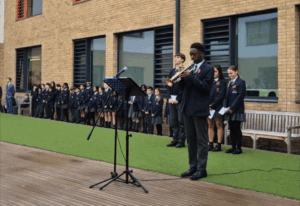 A student in school uniform plays a trumpet during an outdoor ceremony. A line of fellow students stands respectfully behind him against a brick school building.