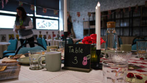A table setting for a Remembrance Day tea, featuring a small chalkboard sign reading "Table 3." The table includes a gray mug, water glasses, tall white candles, and a small arrangement of red poppies. In the softly lit background, a person stands near a window in a room decorated with commemorative banners