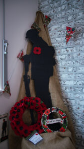 Remembrance Day display featuring a large black silhouette of a soldier wearing a red poppy, resting on a hessian backdrop. A traditional red poppy wreath and a smaller "Lest We Forget" wreath are displayed at the base.