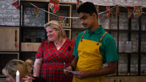 a young man in a green shirt and a yellow apron with suspenders, takes a note or order from a smiling woman wearing a red tartan dress and a poppy pin on her chest. They are standing in a decorated room with shelving units and a white brick-patterned wall. Above them, a banner with the phrase "Lest We Forget" is visible, indicating a Remembrance Day event.