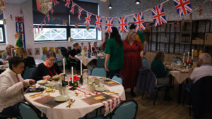 A wide shot of a room full of people attending a Remembrance Day commemorative tea. The room is heavily decorated with Union Jack flags and banners, with a large window in the background. Several round tables are set with tablecloths, light gray mugs, plates, lit candles, and newspaper-themed placemats. Guests are seated and eating. Two people who appear to, one in a red tartan dress and one in a green shirt, are standing in the center-right of the room.