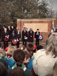 Photo of a Remembrance Day ceremony with a large crowd in the foreground and a group of officials and students holding wreaths at a brick war memorial in the background.