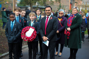Group photo taken outdoors during a Remembrance Day parade, showing students holding a poppy wreath, an adult in a suit, and other attendees in the background.