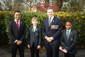 Formal photo of two men in suits, one wearing military medals, standing with two students in school uniform, all wearing poppies.